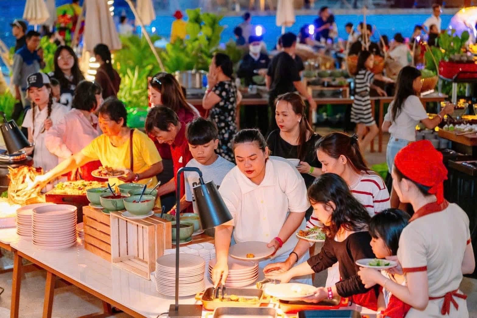 Du khách xếp hàng lấy món tại quầy buffet ngoài trời ban đêm bên hồ bơi trong công viên